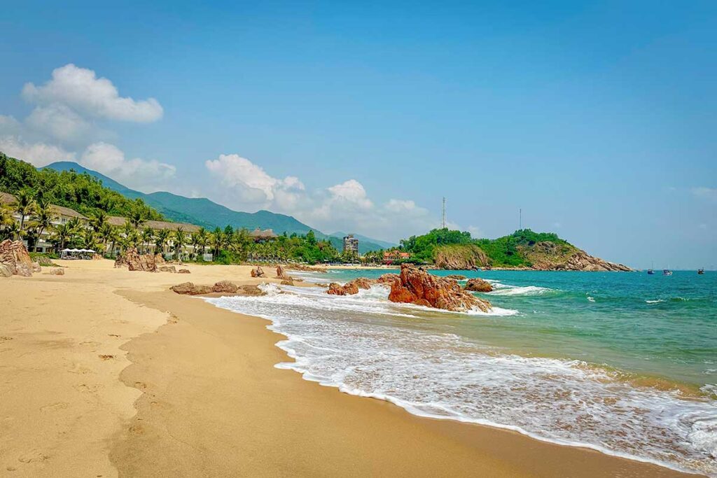 Bai Xep Beach with resorts and mountains in Quy Nhon – Sandy stretch of Bai Xep Beach framed by palm trees, rocky outcrops, and the green mountains of Quy Nhon, with beachfront resorts in the background.