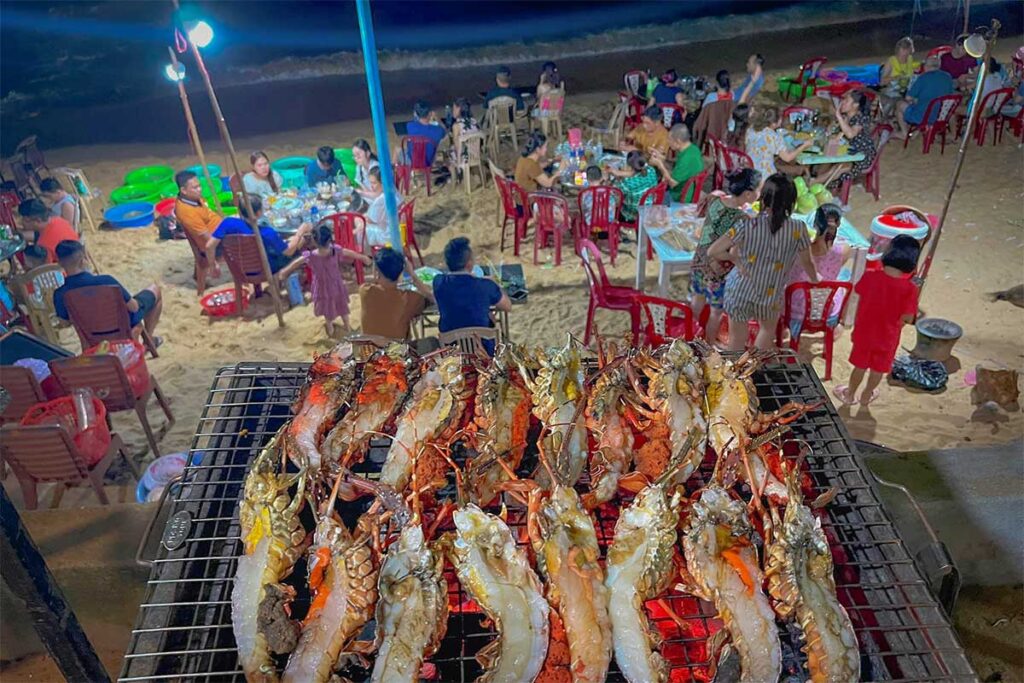 Seafood BBQ on Bai Xep Beach at night – Fresh prawns and lobster grilled on open flames, with plastic chairs and locals dining under bright lights right on the sand.