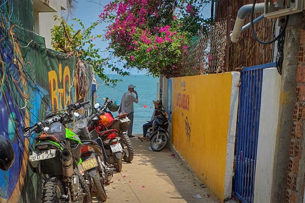 Narrow alley in Bai Xep fishing village leading to the beach, lined with motorbikes, colorful murals, and bougainvillea overlooking the sea in Quy Nhon.