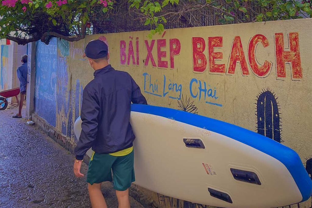 Entrance lane to Bai Xep Beach – Local alleyway with painted sign and traveler carrying a SUP board, leading toward the fishing village beach.