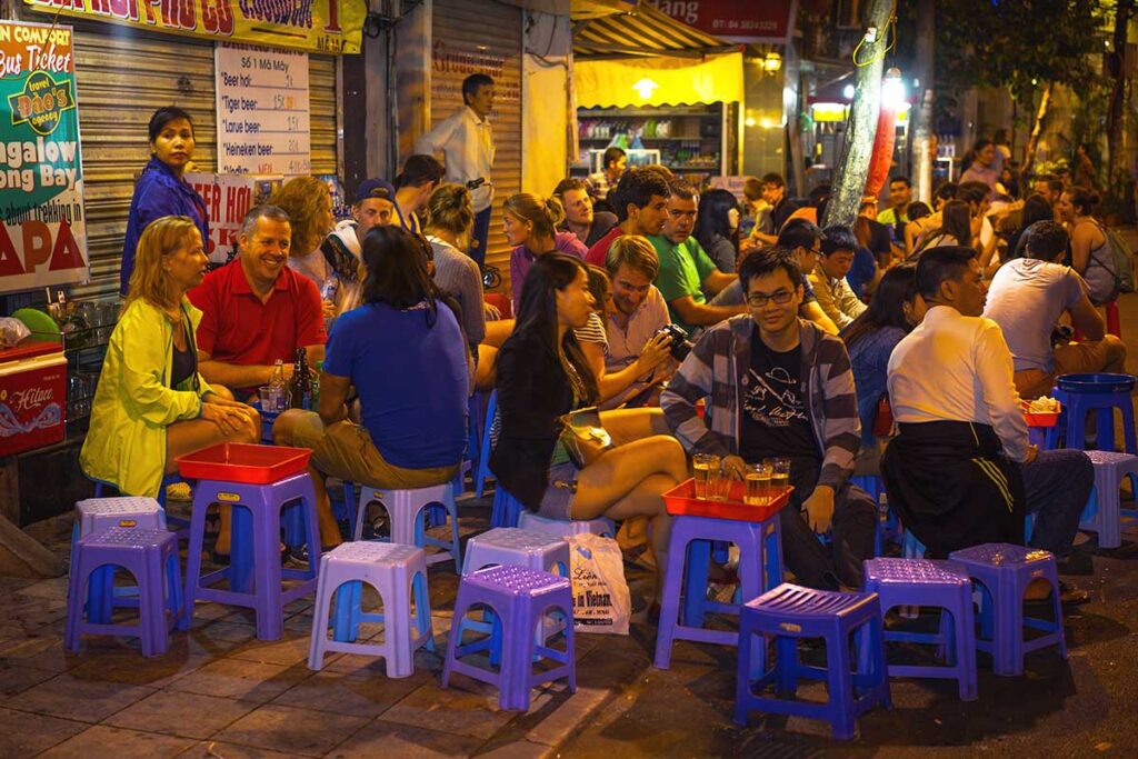 Tourists and locals sitting on plastic stools low to the ground on the street drinking bia hoi at Bia Hoi Junction