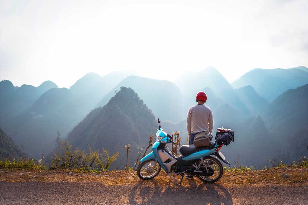 A man standing next to his motorbike on the Ha Giang Loop route, with a view of stunning mountain scenery, one of the best things to do in Vietnam.