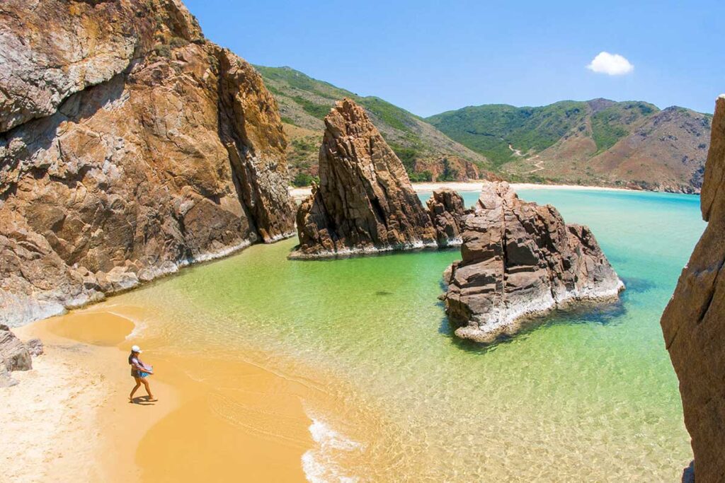 Tourist walking in shallow emerald water between dramatic rock formations at Ky Co Beach in Quy Nhon, Vietnam.
