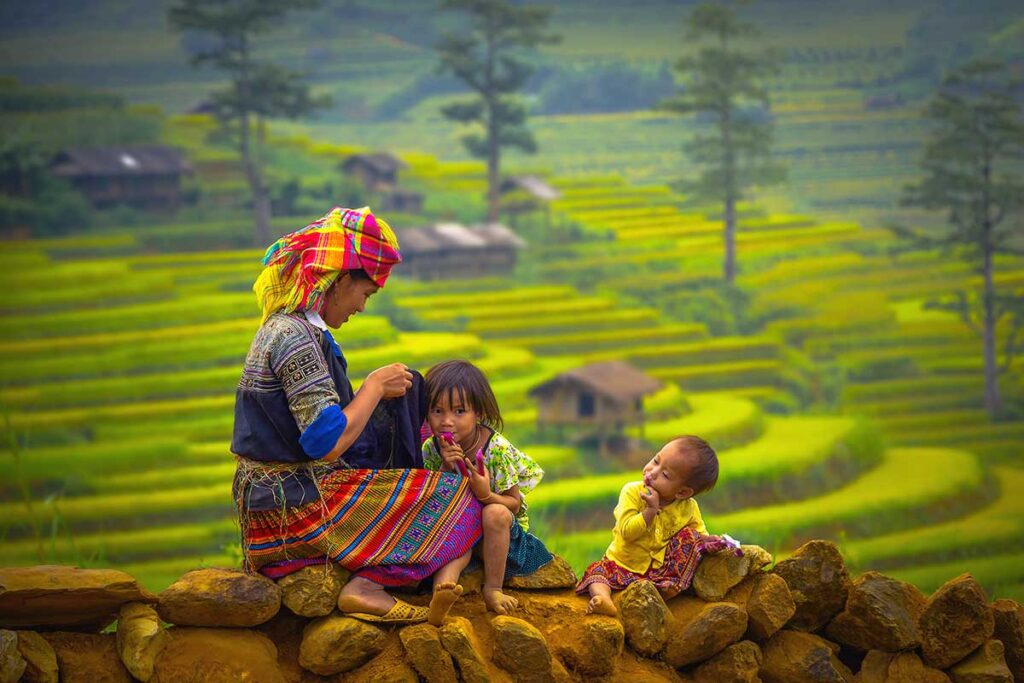 A colorful ethnic minority woman sitting on a stone wall with two children, with terraced rice fields of Sapa in the background.