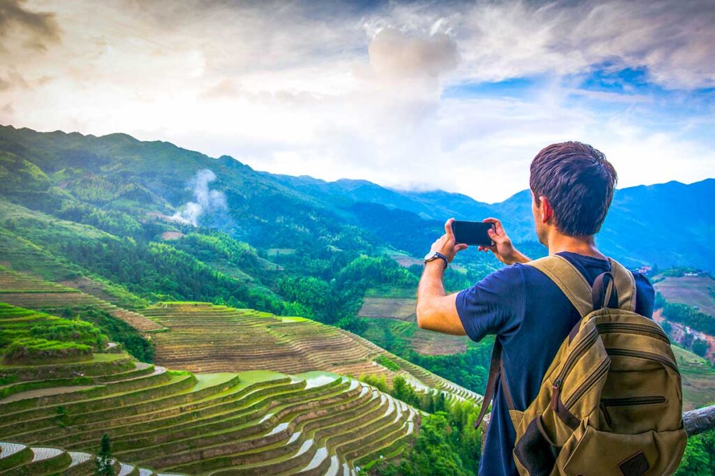 A tourist man making a trekking in Sapa makes a stop wearing a backpack and holding his phone to take a photo of the terraced rice fields.