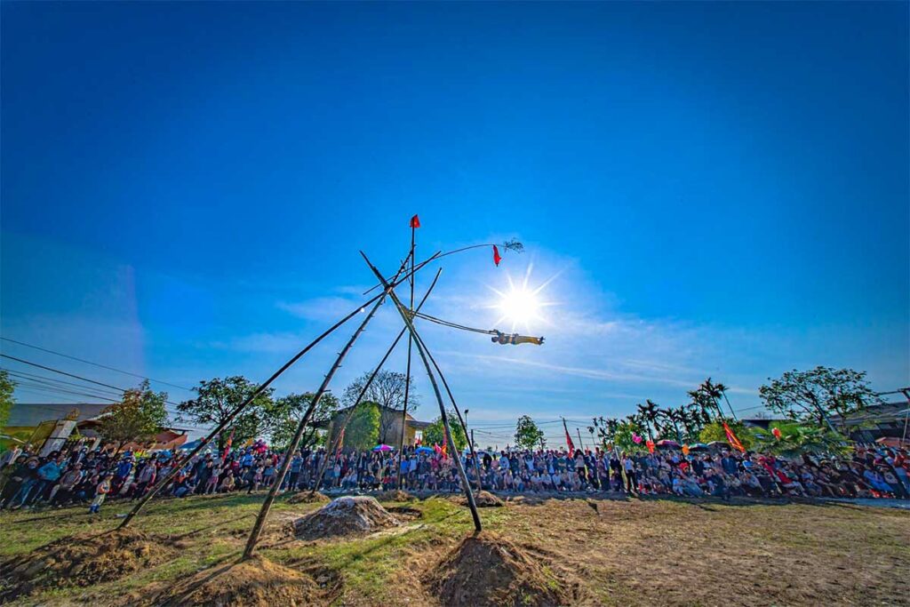 A man sitting on a very large bamboo swing, swinging very high in the air while a large audience around it is watching the game during Du Tien Festival