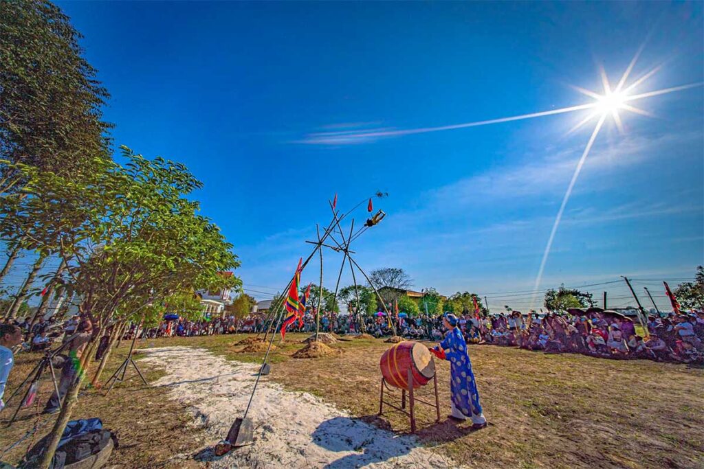 A man in blue traditional clothes is hitting a large drum with in the background a giant bamboo swing that is used for Du Tien Festival in Hue