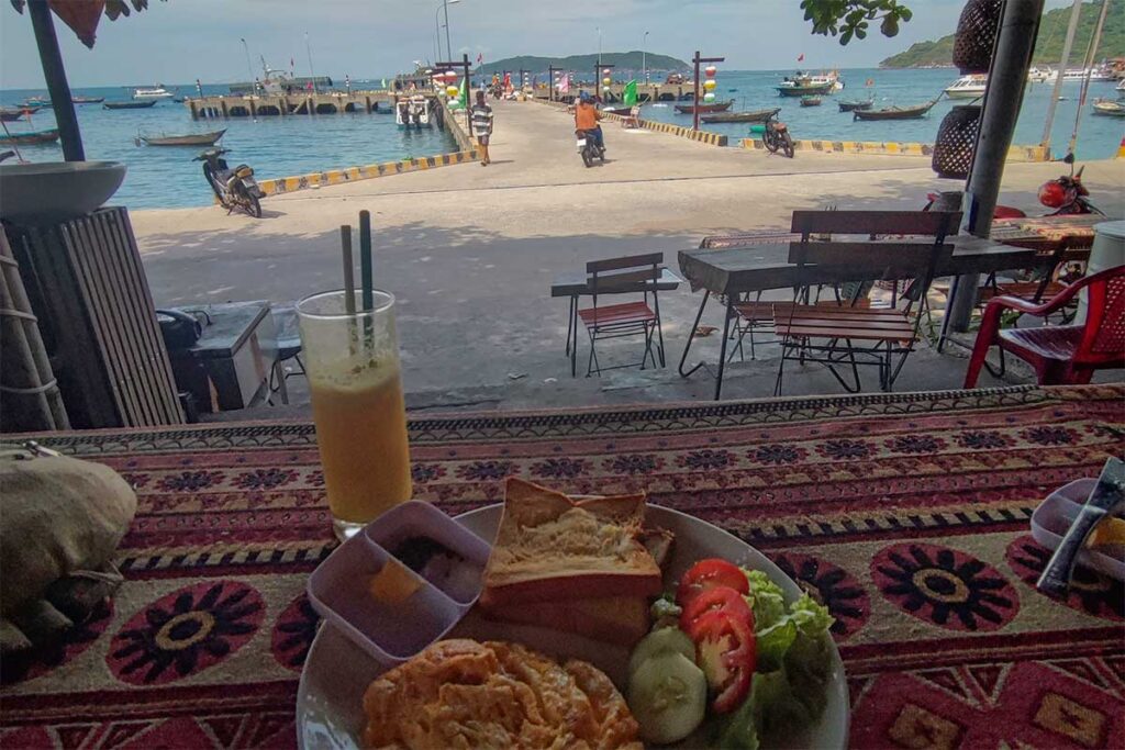 Beachfront café on Cham Island serving local breakfast with sea view over the pier and anchored fishing boats.