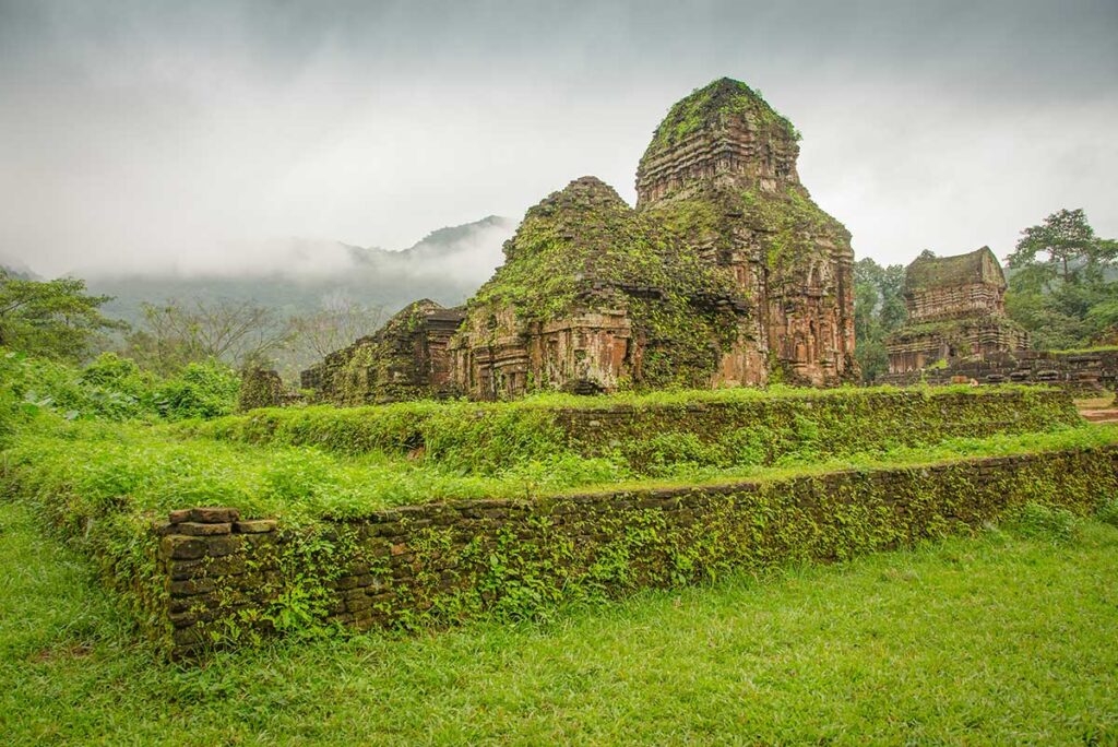 Cham temple structures at My Son Sanctuary with moss-covered walls and mountain mist in the background.”