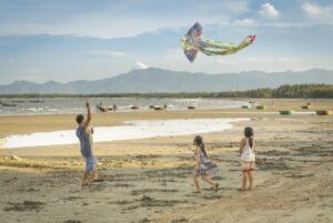 A father and children fly a colorful kite on the quiet beach of Tam Hai Island, with round fishing boats and distant mountains in view.