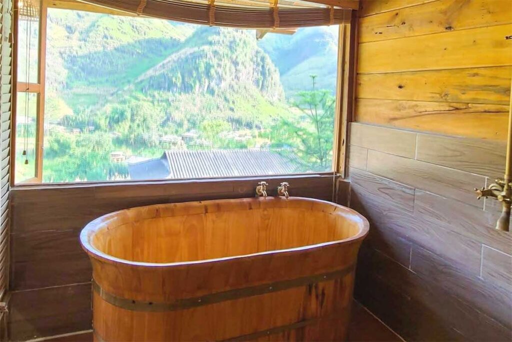 Mountain-view wooden bath in a Du Gia homestay – A rustic wooden bathtub inside a homestay in Du Gia, offering a relaxing soak with a window view of the stunning mountain landscape.