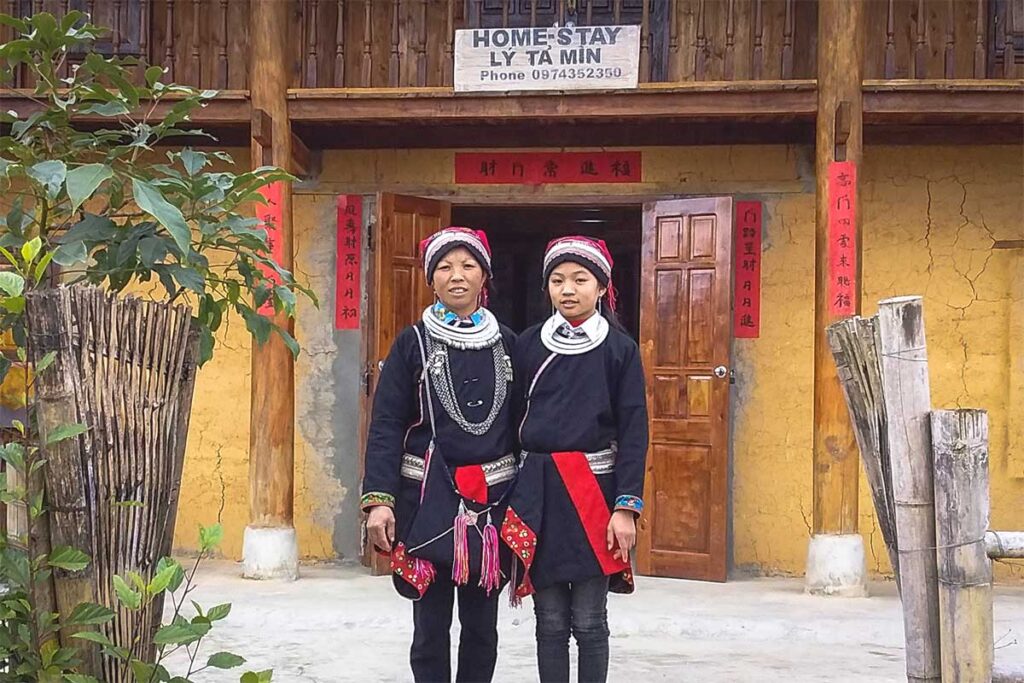 Two Dao ethnic women in traditional red and black clothing standing in front of a yellow clay house used as a homestay in Nam Dam Village.