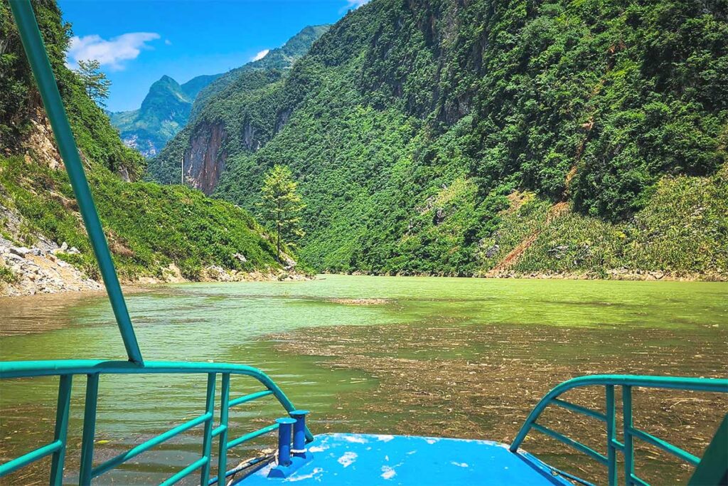 A boat sailing through the Nho Que River at Khau Vai, passing between lush green mountains and dense forests.