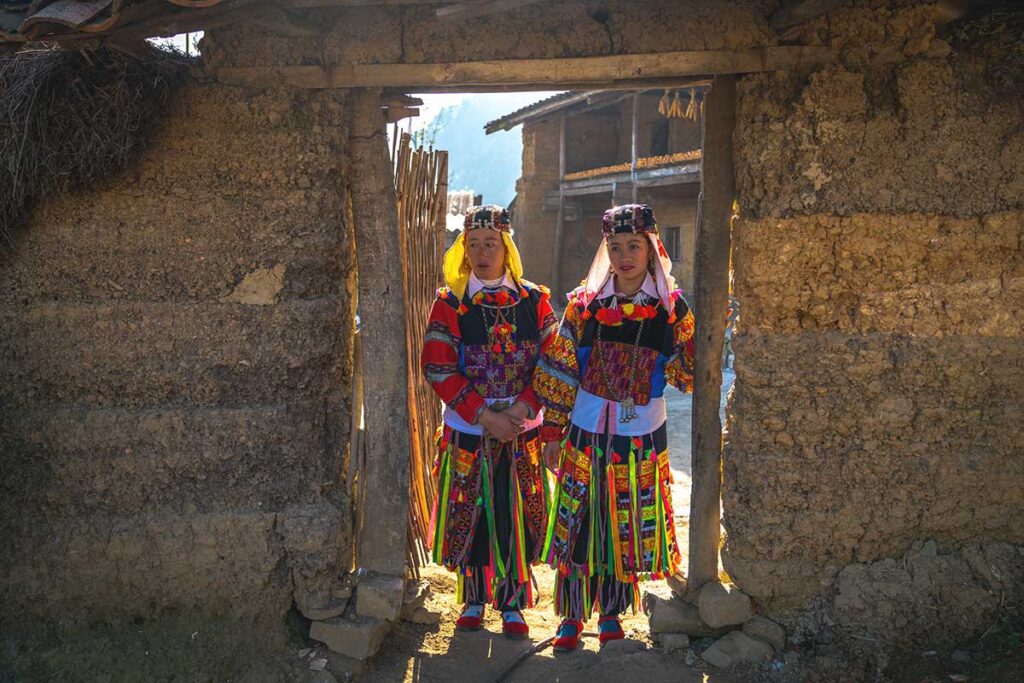 Lo Lo women at their home entrance – Two Lo Lo ethnic women in colorful traditional clothing standing at the entrance of their clay house, giving a glimpse into local life.