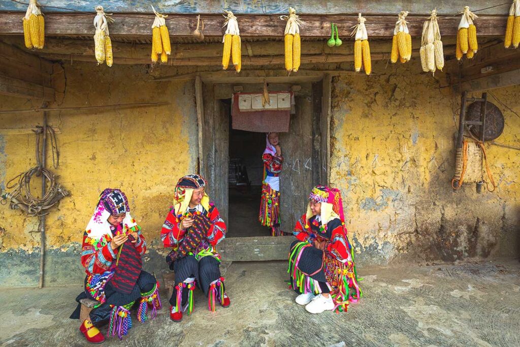 Lo Lo women in traditional attire – Three Lo Lo ethnic women in their vibrant, multicolored outfits sitting in front of a traditional yellow clay house in Lo Lo Chai Village.
