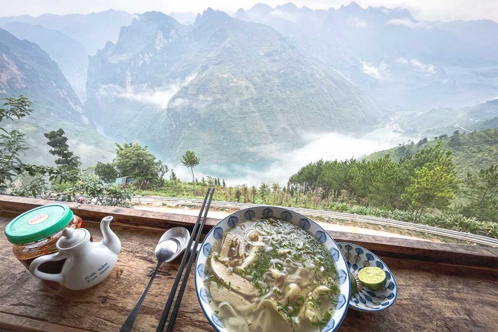 A bowl of steaming pho placed on a table with a breathtaking view of Ma Pi Leng Pass from Ma Pi Leng Eco Lodge.