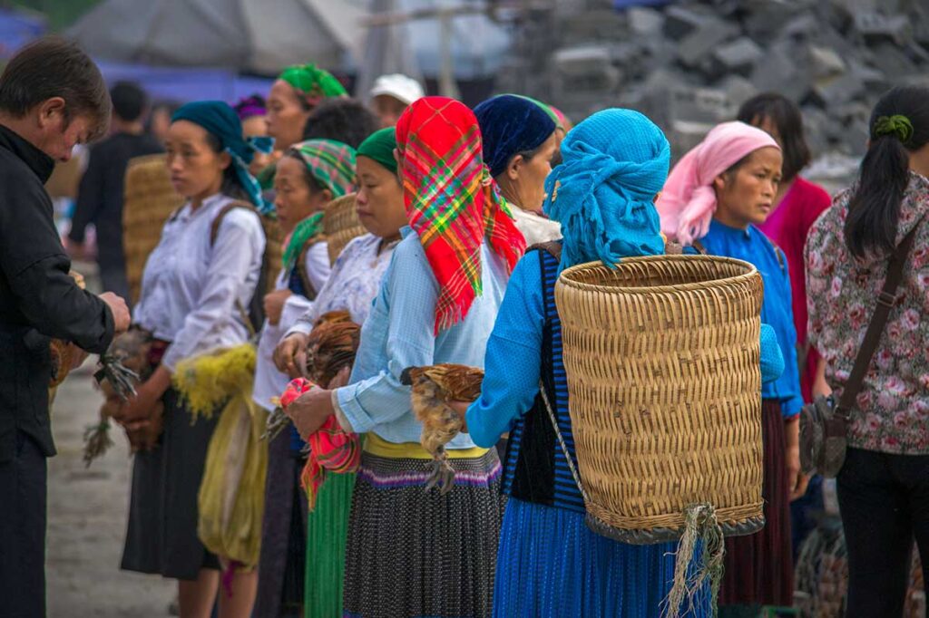 A group of ethnic women in traditional clothing at Meo Vac Market, wearing colorful scarves and holding chickens.