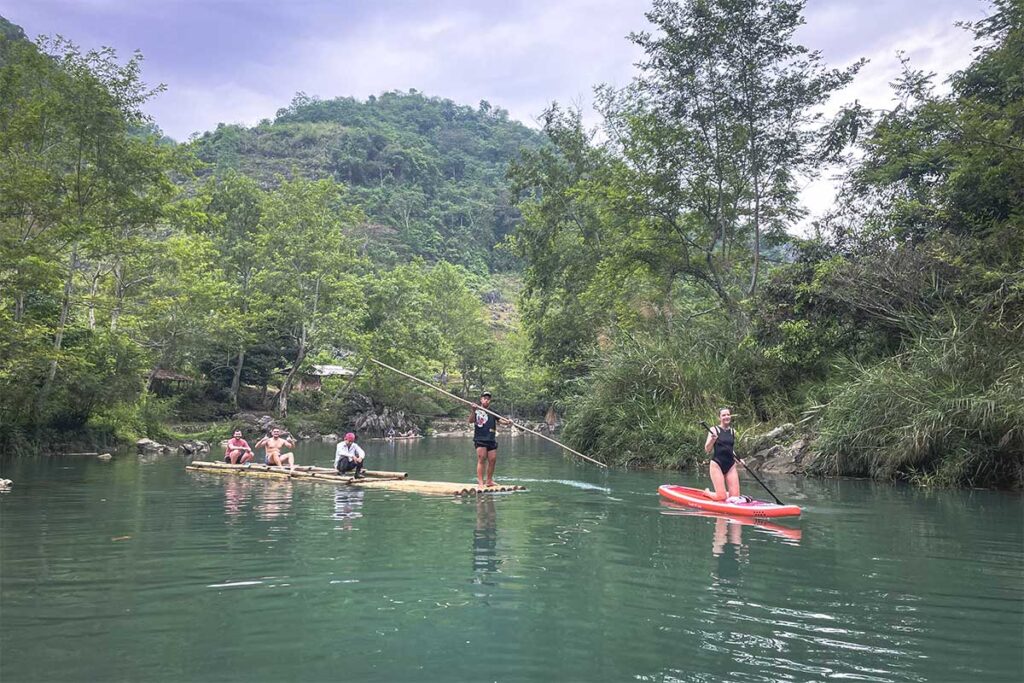 Bamboo rafting at Tham Lin Cave – Three tourists on a bamboo raft, guided by a local using a long pole, while another traveler paddles on a stand-up board through the scenic forested stream at Tham Lin Cave.