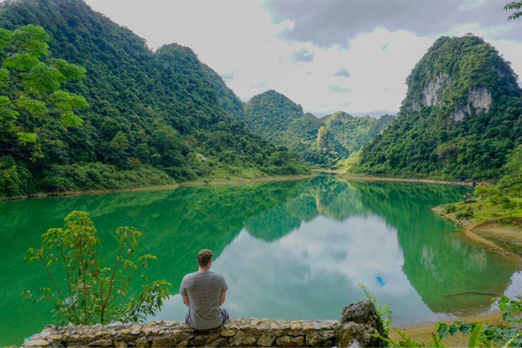 A man sitting on a small stone wall overlooking Thang Hen Lake in Cao Bang, reflecting the surrounding mountains in the lake