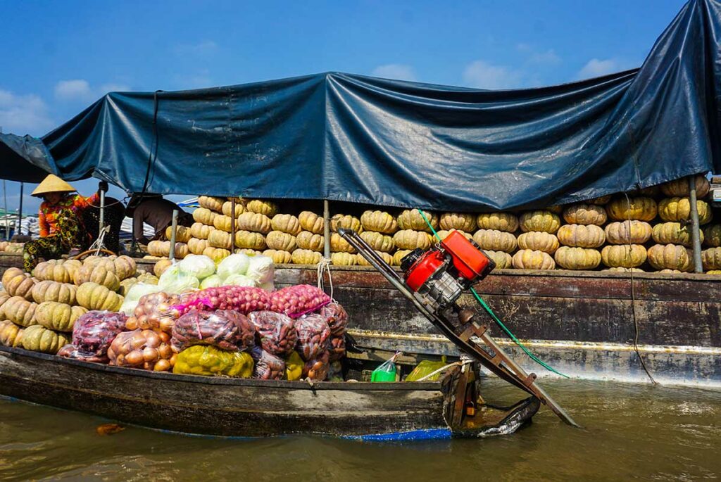 Large wholesale boat stacked with pumpkins and onions at Cai Rang Floating Market, with a smaller sampan pulling up alongside.
