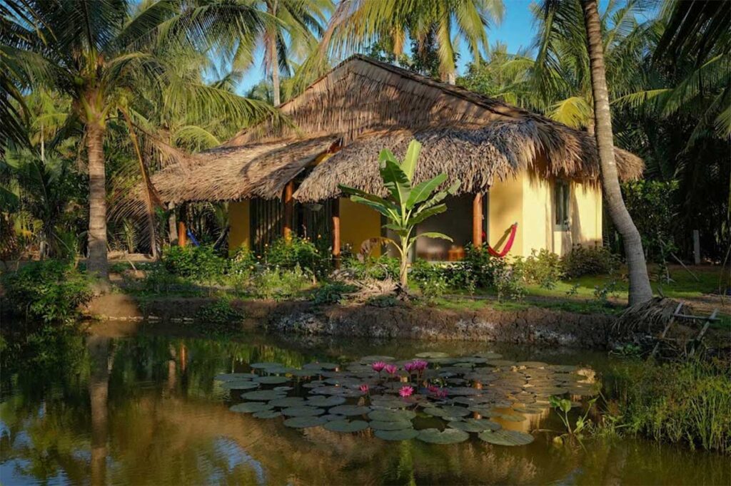 Traditional homestay in Ben Tre with thatched roof, hammock, and lotus pond surrounded by coconut palms in the Mekong Delta, Vietnam.
