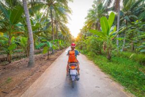 Traveler riding a scooter through coconut palm and banana tree paths in Ben Tre countryside, Mekong Delta, Vietnam.