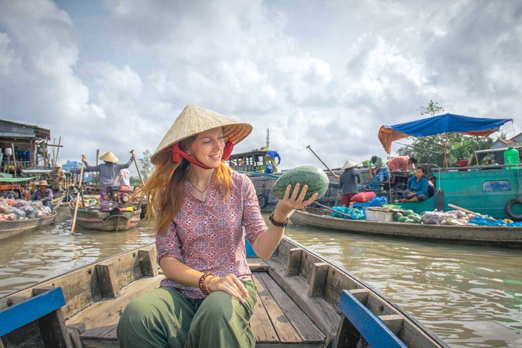 Traveler wearing a conical hat holding a watermelon on a boat ride at Cai Rang Floating Market in Can Tho.