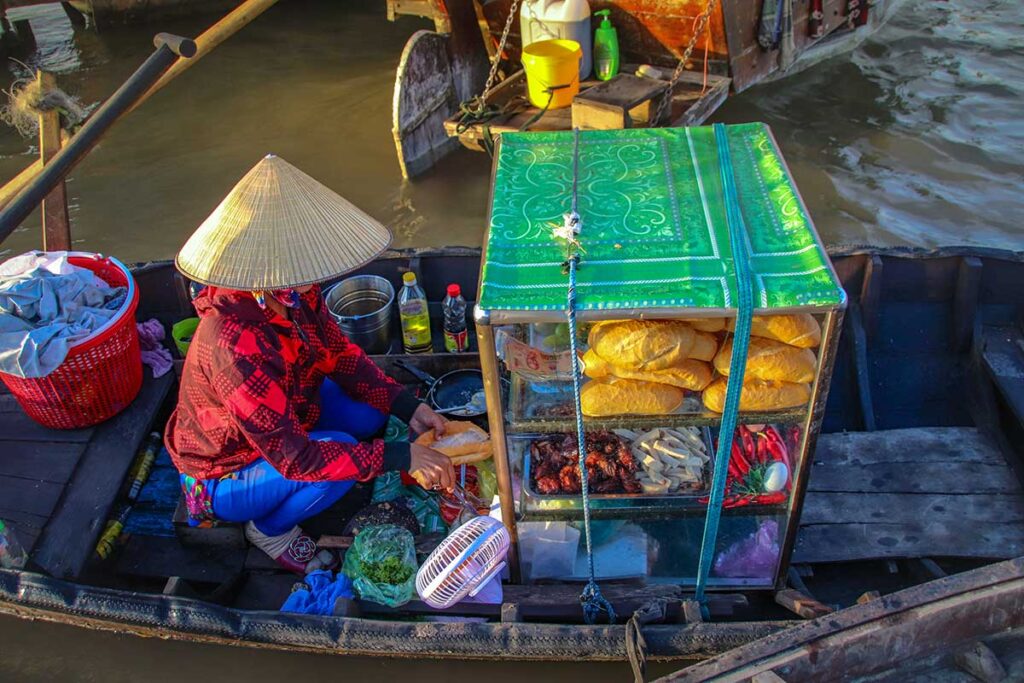 Vendor preparing fresh bánh mì sandwiches from a small food boat at Cai Rang Floating Market, with loaves stacked in a glass case.