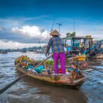 Sampan loaded with pineapples being rowed through Cai Rang Floating Market at sunrise, surrounded by larger cargo boats.