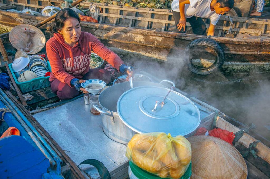 Local woman preparing steaming noodle soup from a floating kitchen boat at Cai Rang Floating Market, serving breakfast to visitors.