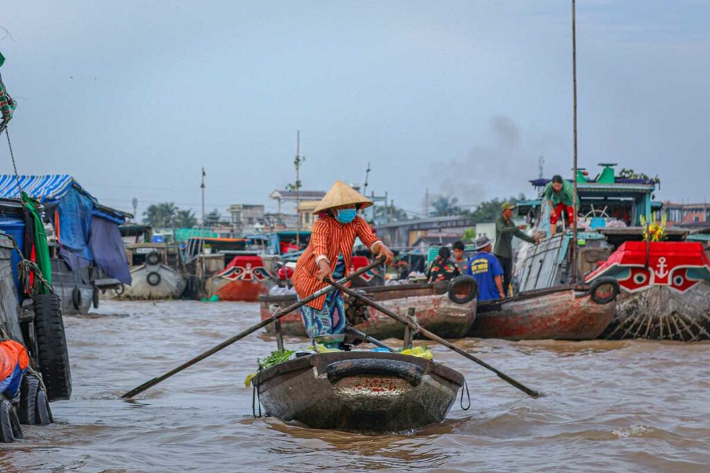 Local woman rowing a small sampan through the busy waterways of Cai Rang Floating Market, surrounded by larger trading boats.