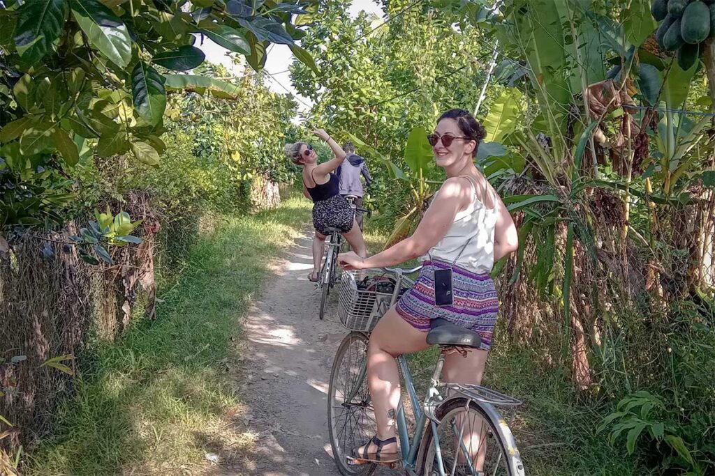 Tourists riding bicycles along narrow rural trails surrounded by fruit orchards in Can Tho countryside.