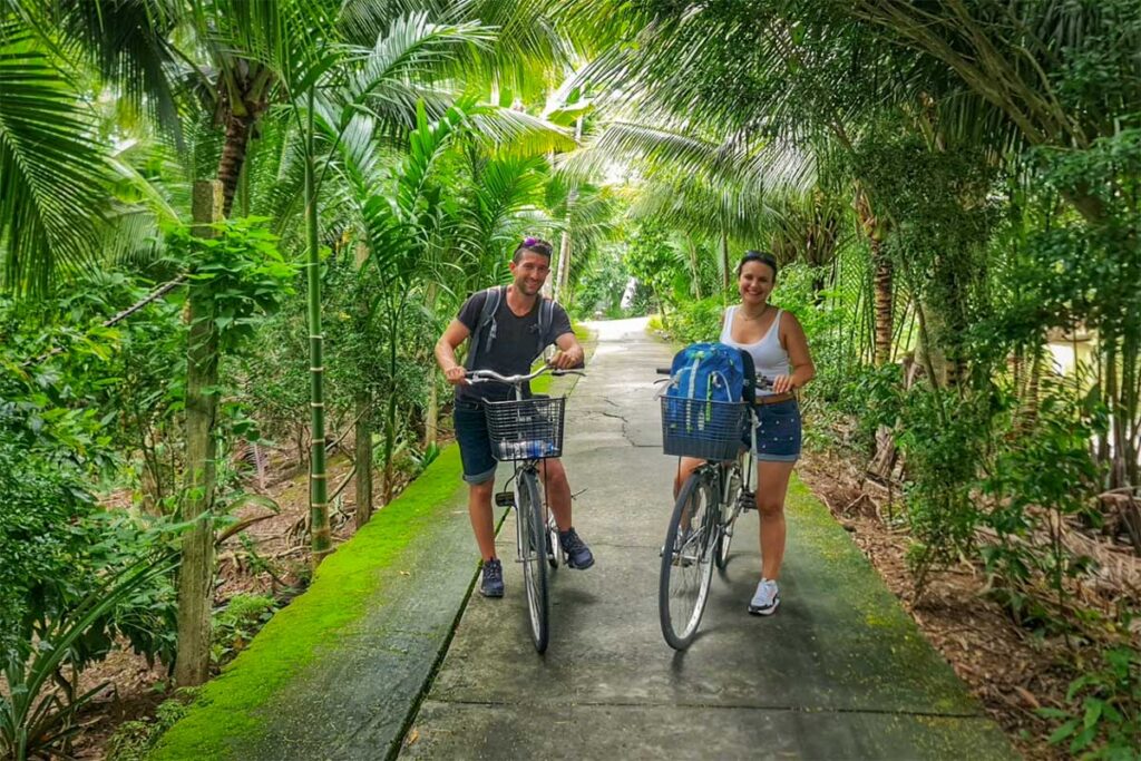 Cycling tour through Can Tho village roads – Couple with bicycles on a narrow concrete path, surrounded by palm trees and lush greenery.