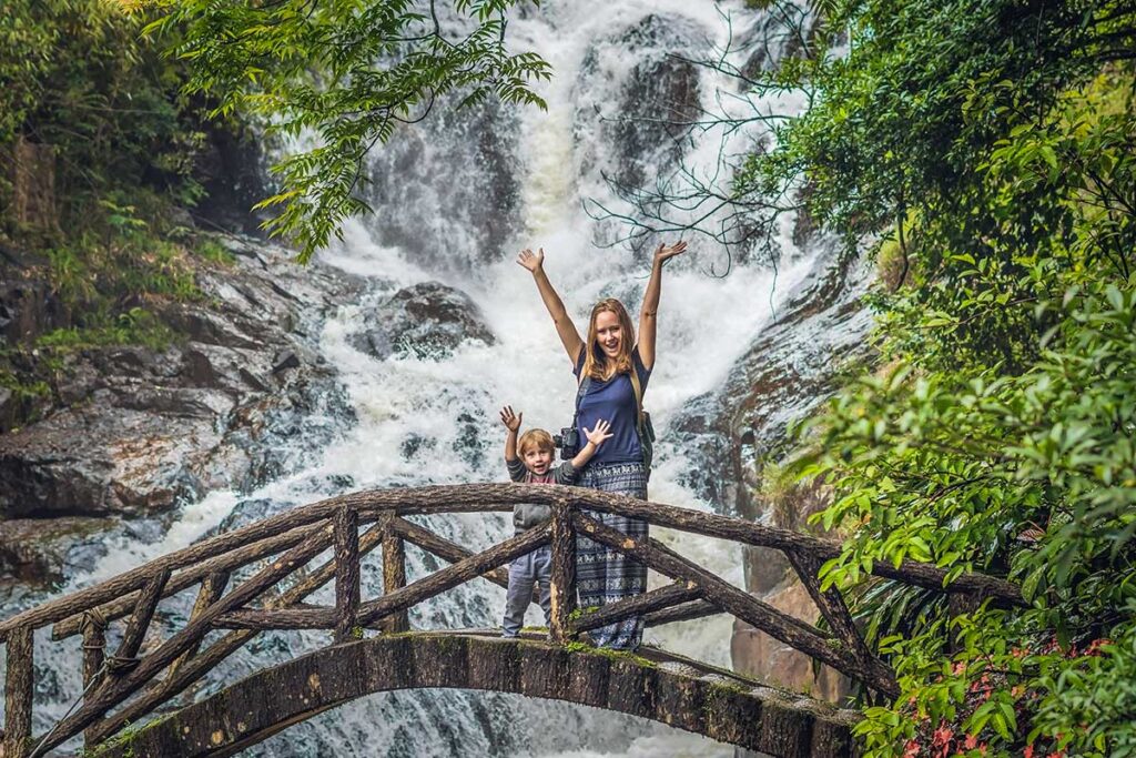 A mother and her young son stand on a small wooden bridge with their arms raised in front of the rushing Datanla Waterfall near Dalat, a fun nature stop on a family holiday in Vietnam with kids.