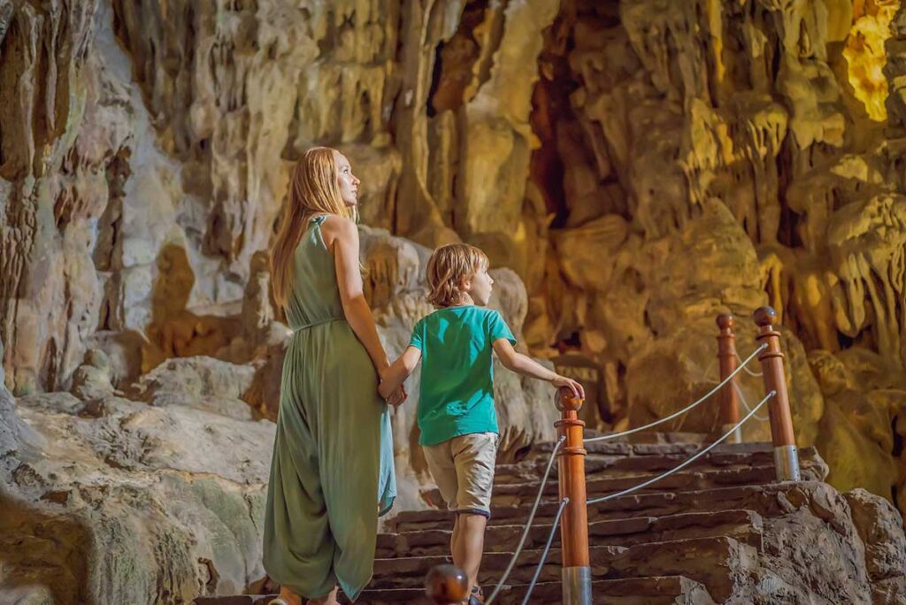 A mother and her son walk up the lit stone steps inside a large cave in Halong Bay, looking up at the rock formations, an easy and interesting stop when traveling with children.