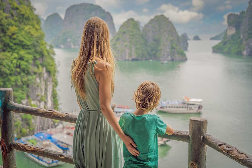 A mother and her young son stand at a wooden railing on a family holiday in Vietnam with kids, looking out over the limestone karsts and tour boats of Halong Bay.