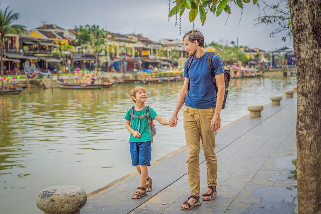 A father and son hold hands while walking along the riverfront in Hoi An ancient town, with colorful boats and old yellow houses behind them, a relaxed stop when traveling with children in Vietnam.