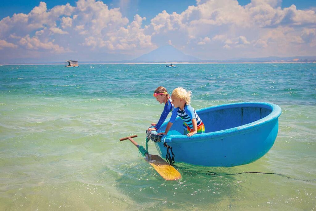 Two children sit in a round blue basket boat in the clear shallow water off Nha Trang, with a mountain in the distance, an easy way to enjoy beach time on a family holiday in Vietnam with kids.