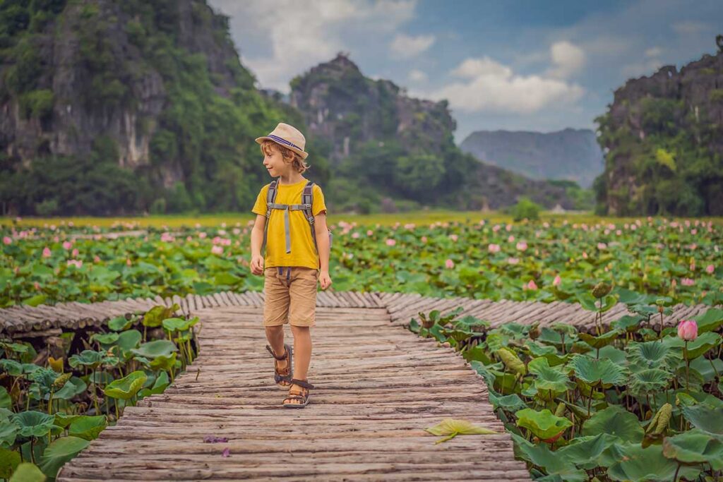 A boy with a backpack walks along a wooden path crossing a wide lotus pond in Ninh Binh, framed by karst mountains, a peaceful scene on a family holiday in Vietnam with kids.
