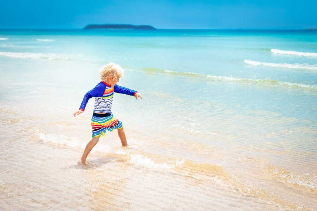 A young boy in a colorful rash guard runs through the shallow water on a clear, calm beach in Phu Quoc, ideal for relaxed beach time on a family holiday in Vietnam with kids.