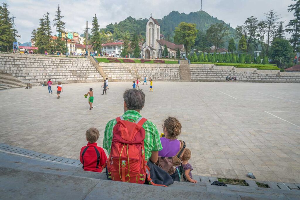 A family with young children sits on the steps of the main square in Sapa town, watching local kids play football in front of the old stone church, an easy and relaxed spot for families.