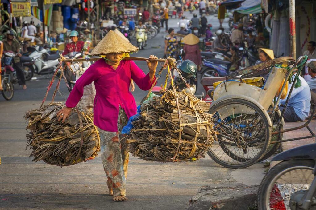 Local woman in My Tho carrying firewood with a traditional shoulder pole at the busy market streets of the Mekong Delta.