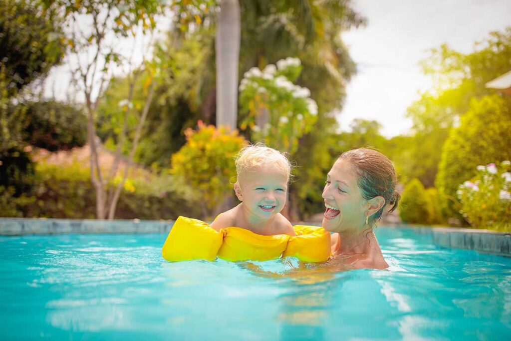A laughing mother and her toddler in yellow arm floats play together in a swimming pool surrounded by greenery, the kind of family friendly accommodation many parents look for in Vietnam.