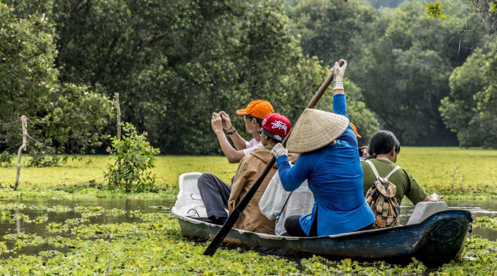 Travelers in a wooden boat taking photos while paddling through duckweed-filled waters of Tra Su Cajuput Forest.