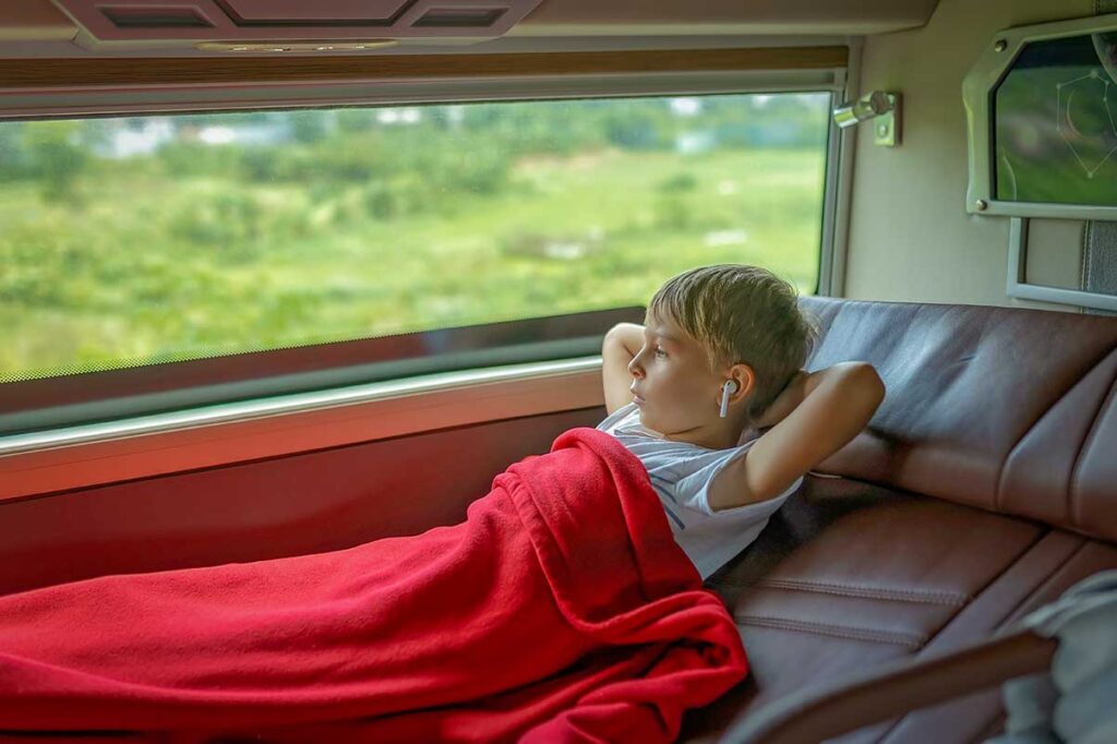 A relaxed young boy lies on a padded berth under a red blanket and looks out the window at the passing green countryside, a calm way to break up long journeys when traveling with children in Vietnam.