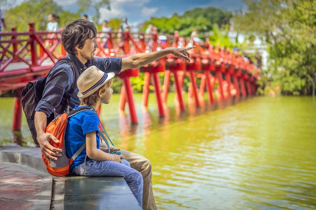 A father points across the water toward the red Huc Bridge at Hoan Kiem Lake while his son sits beside him, an easy and central spot for families exploring Hanoi with kids.