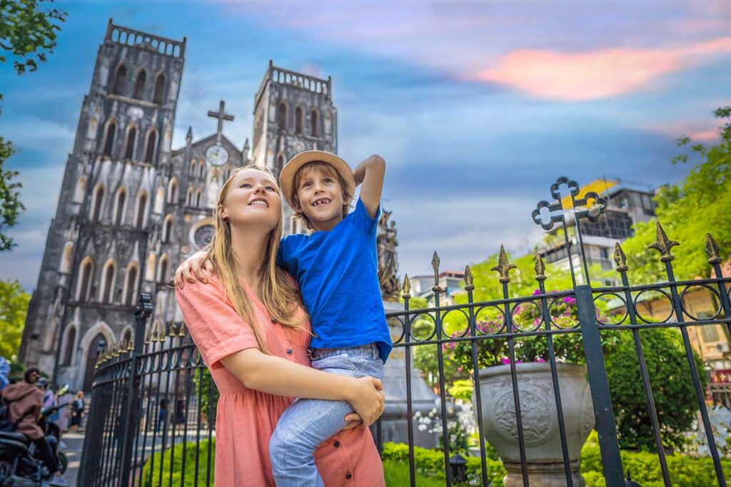 A mother holds her smiling son in front of the twin towers of St Joseph Cathedral in Hanoi at dusk, a quick photo stop while traveling with children in the old quarter.