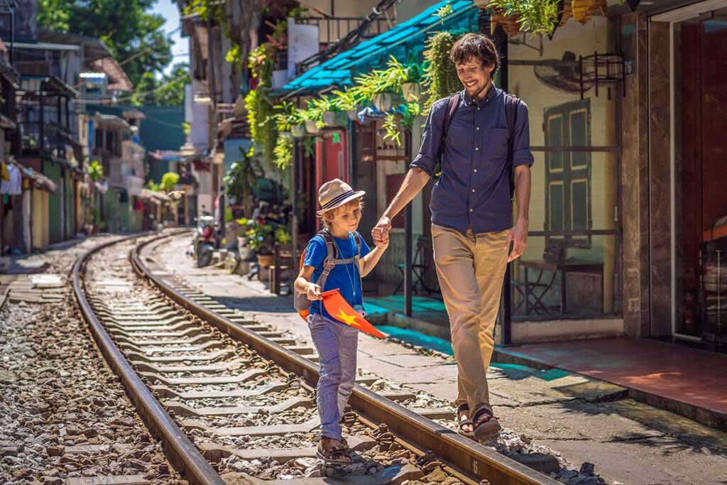 A father and son walk hand in hand along the narrow tracks of Hanoi train street, lined with cafes and old houses, a memorable but careful stop when visiting Vietnam with kids.