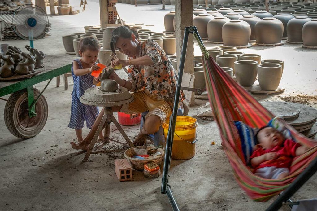 Family pottery workshop in Vinh Long – Mother and child shaping clay figures while a baby rests in a hammock, part of the Mang Thit pottery village tradition.