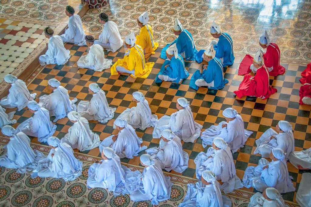 People in white ropes and a few in colored ropes (red, blue and yellow) sitting inside the Cao Dai Holy See Temple in Tay Ninh during praying times